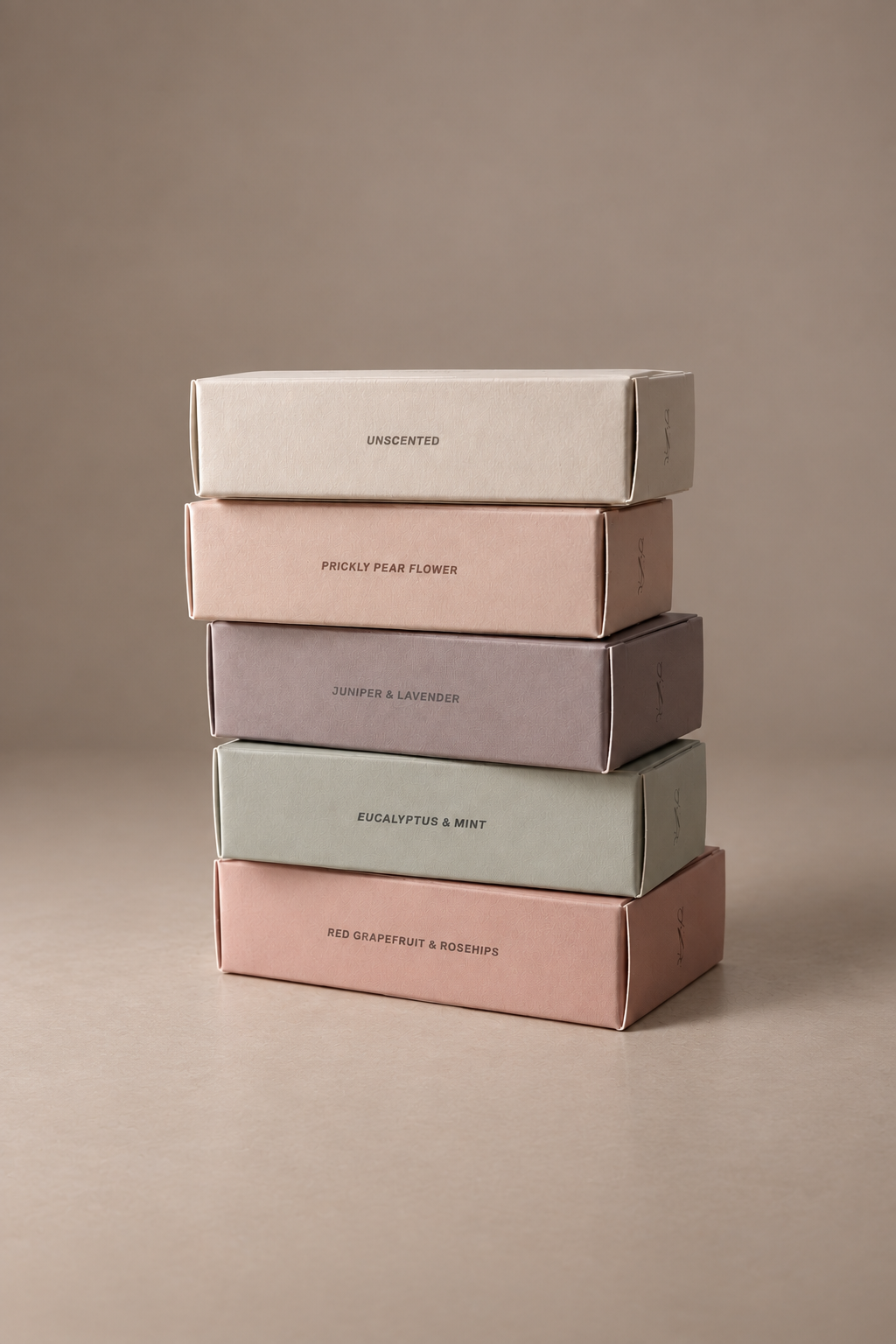 A stack of five minimalist product boxes in muted pastel colors, including cream, blush pink, dusty lavender, sage green, and dusty rose, set against a solid taupe background.