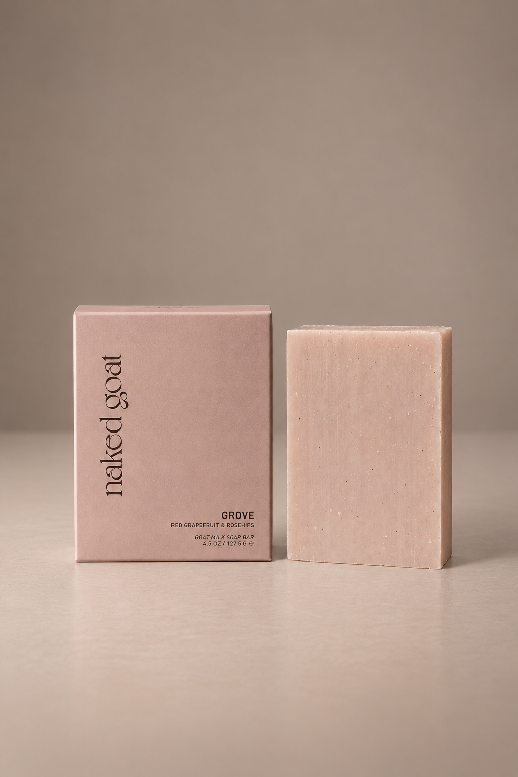 A pale blush pink rectangular bar of soap stands next to its matching pink packaging box on a neutral beige surface and background. The box features the brand name 'naked goat' and the scent name 'GROVE'.