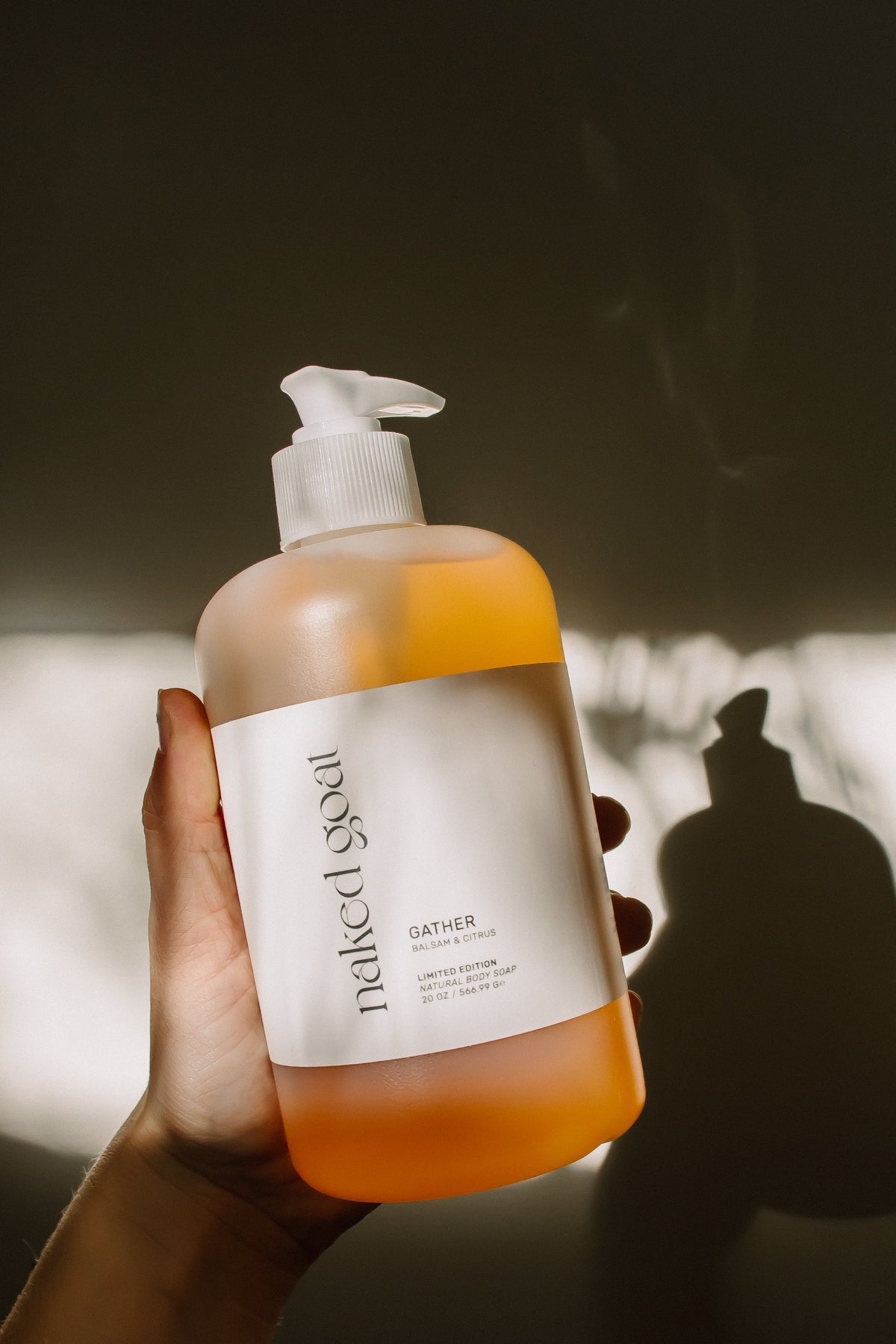 A hand holds a bottle of Naked Goat Gather Hand & Body Soap, showing the orange liquid inside a frosted bottle with a white label and pump dispenser, set against a dark background with dramatic shadows.