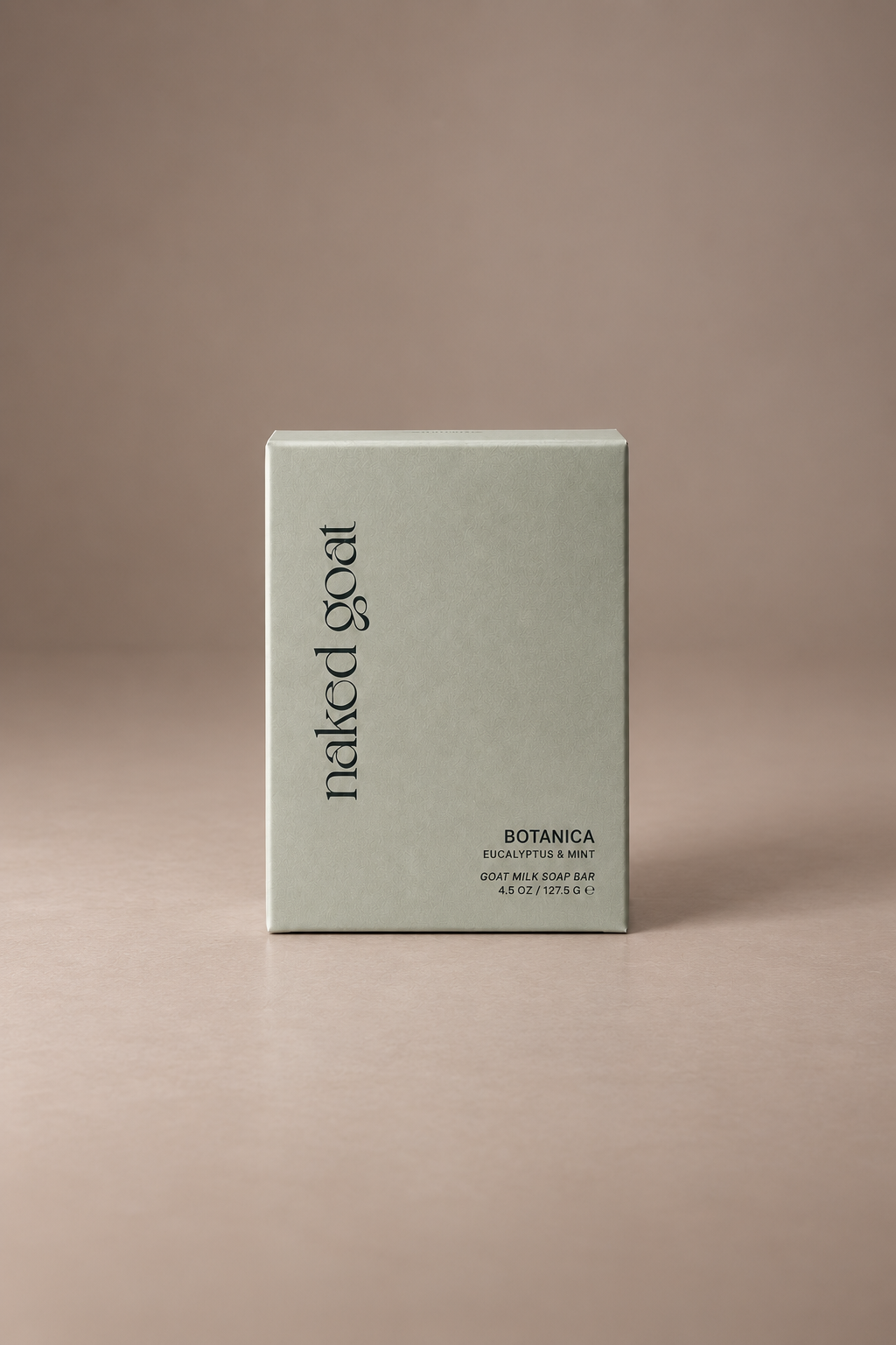 A minimalist light sage green rectangular box for Naked Goat Botanica bar soap, standing upright on a matching beige surface against a solid taupe background.
