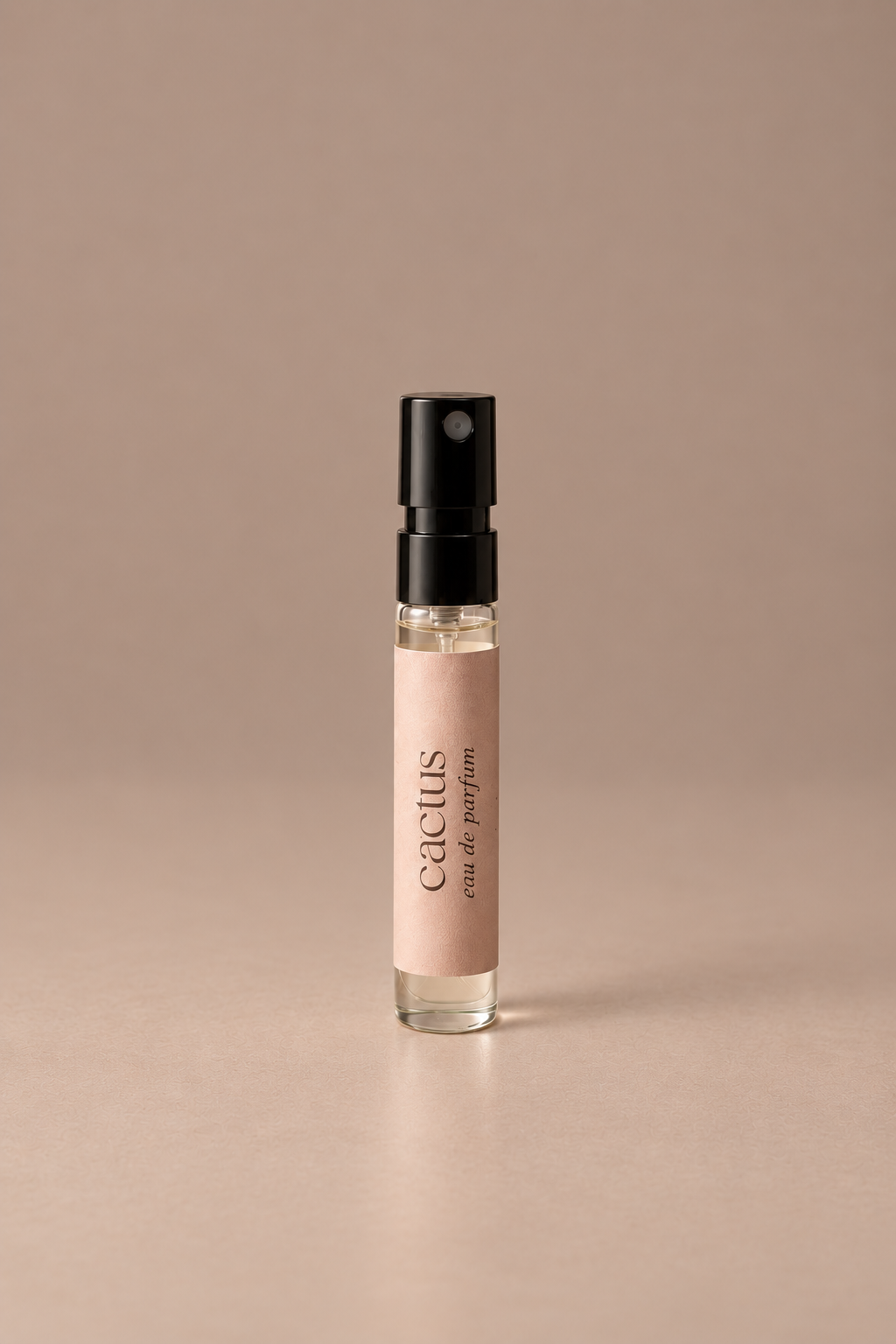 A small cylindrical perfume atomizer bottle with a black spray top and a pale blush pink label reading 'cactus eau de parfum', centered on a warm beige background.