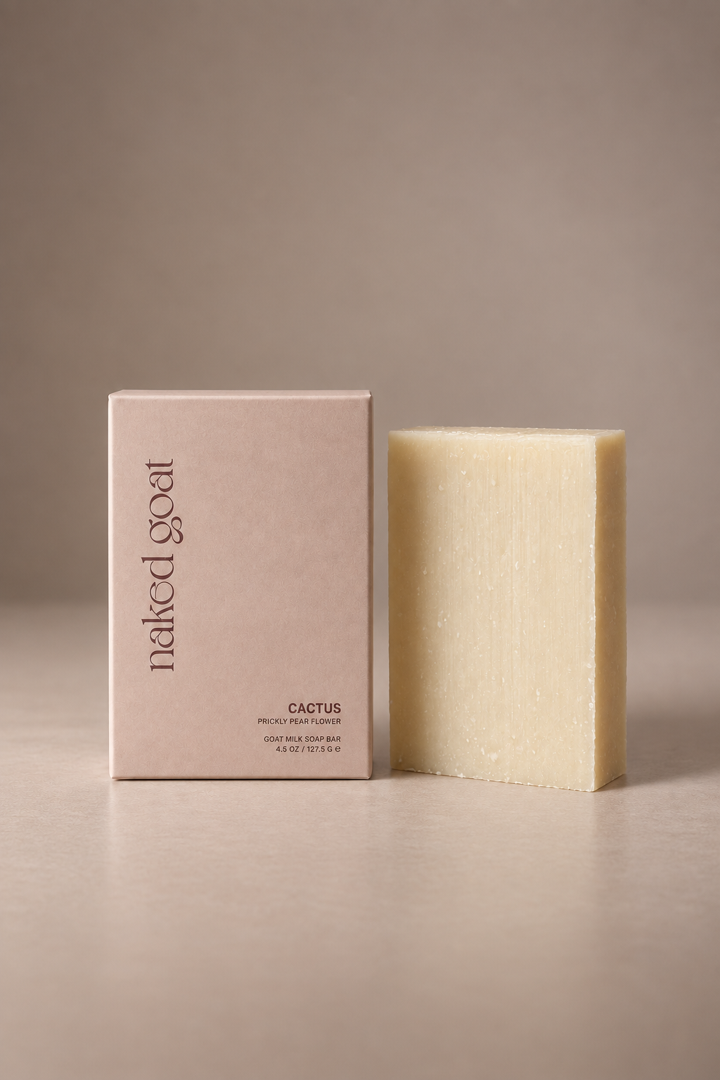 A pale cream-colored soap bar standing next to its textured blush pink packaging box, which features the brand name 'naked goat' and the scent 'CACTUS'. The background is a clean, neutral warm beige studio setting.