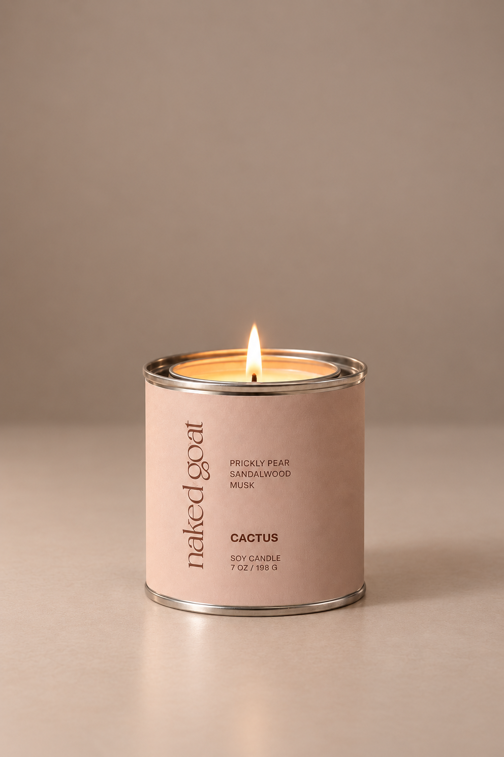 A lit soy candle in a matte blush pink tin with silver metal rims, featuring the brand name 'naked goat' and scent 'Cactus' on the front label, set against a warm beige background.