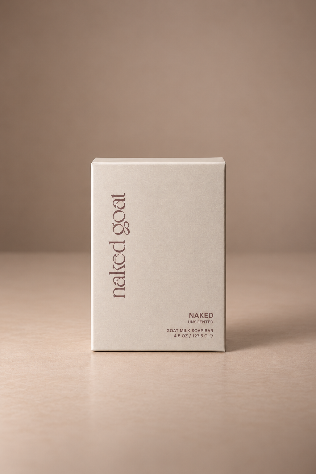 A minimalist off-white rectangular box for Naked Goat NAKED goat milk soap bar, standing upright on a matching beige surface against a neutral beige background.