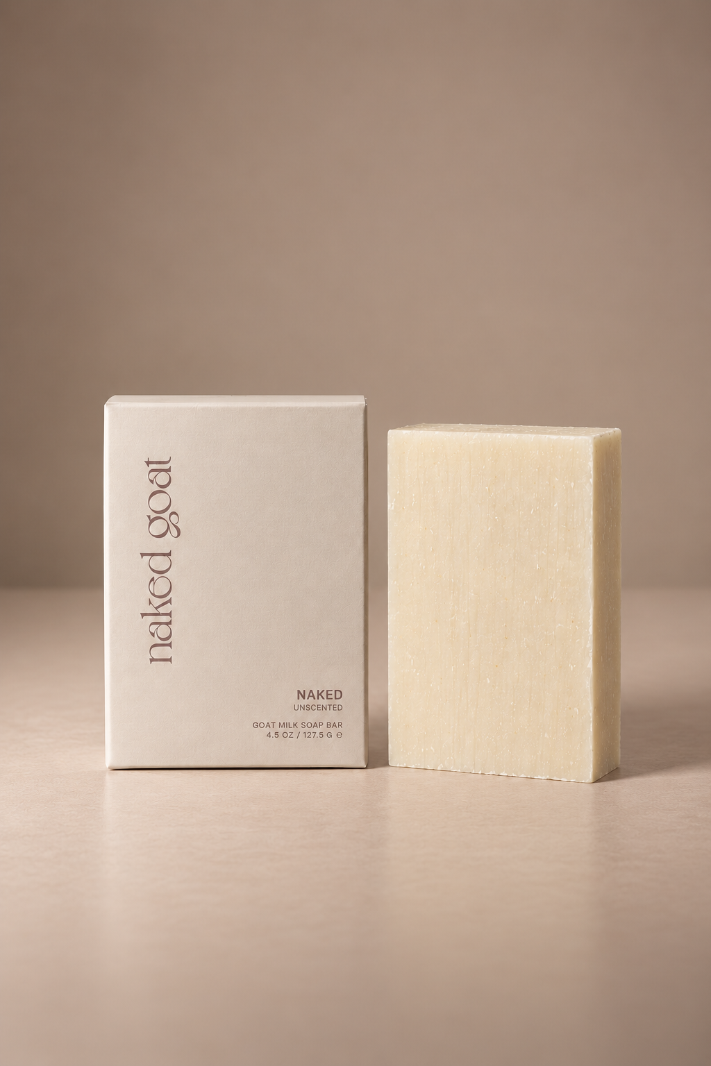 A rectangular bar of natural soap standing next to its minimalist cream-colored packaging box on a neutral beige surface and background. The box features the text 'naked goat' and 'NAKED' in a clean, modern font.