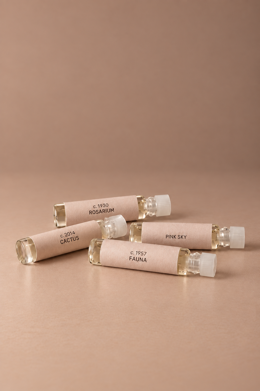 Four small clear glass perfume sample vials with pale blush pink paper labels, arranged on a warm beige background. The labels show different scent names: ROSARIUM, CACTUS, PINK SKY, and FAUNA.